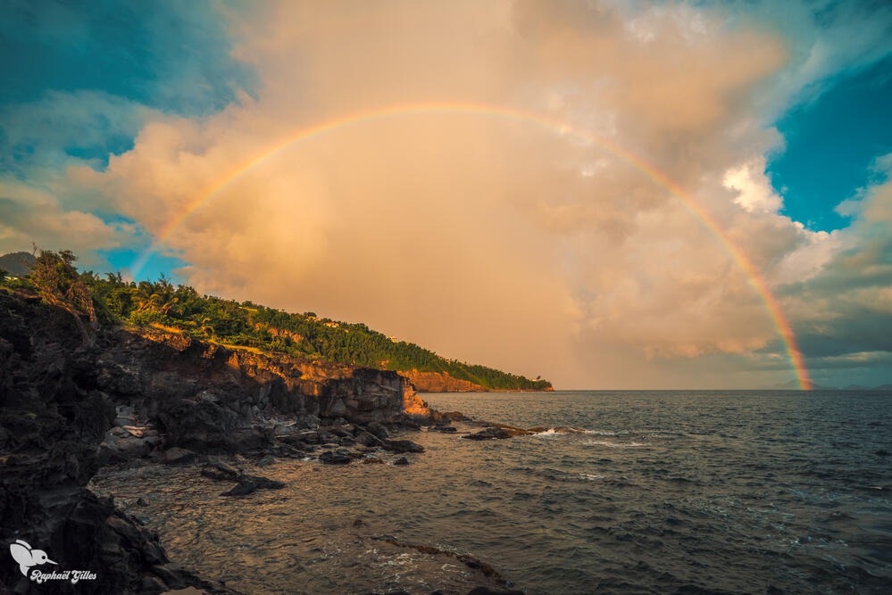 Un arc en ciel pendant un coucher de soleil, la mer en premier plan, les terres de Guadeloupe en fond.
