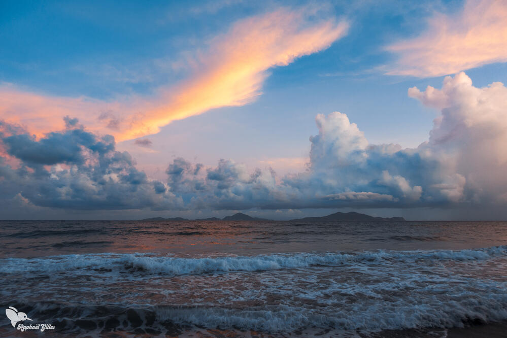 Un coucher de soleil sur la mer des Caraïbes.
Des nuages blancs dans le ciel.
Les îles des Saintes sur l’horizon.