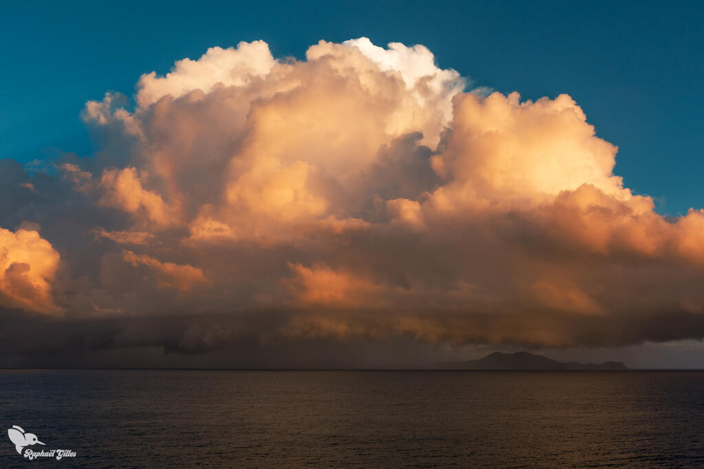 Vue sur la mer. Un nuage de pluie s’abat sur des îles à l’horizon. Les couleurs chaudes du soleil couchant se reflétant dessus.