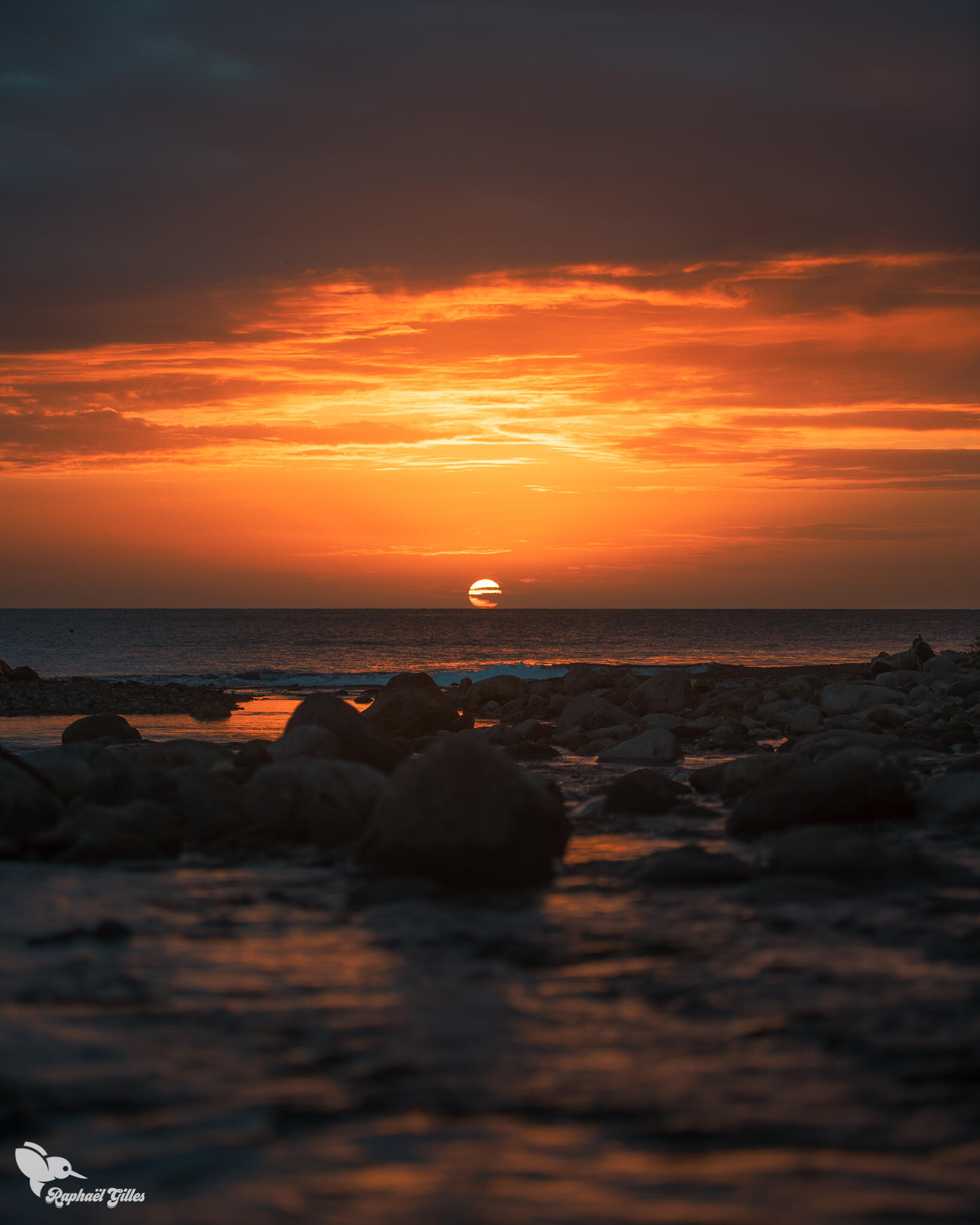 Une rivière se jette dans la mer face à un coucher de soleil dans des tons très orangés.