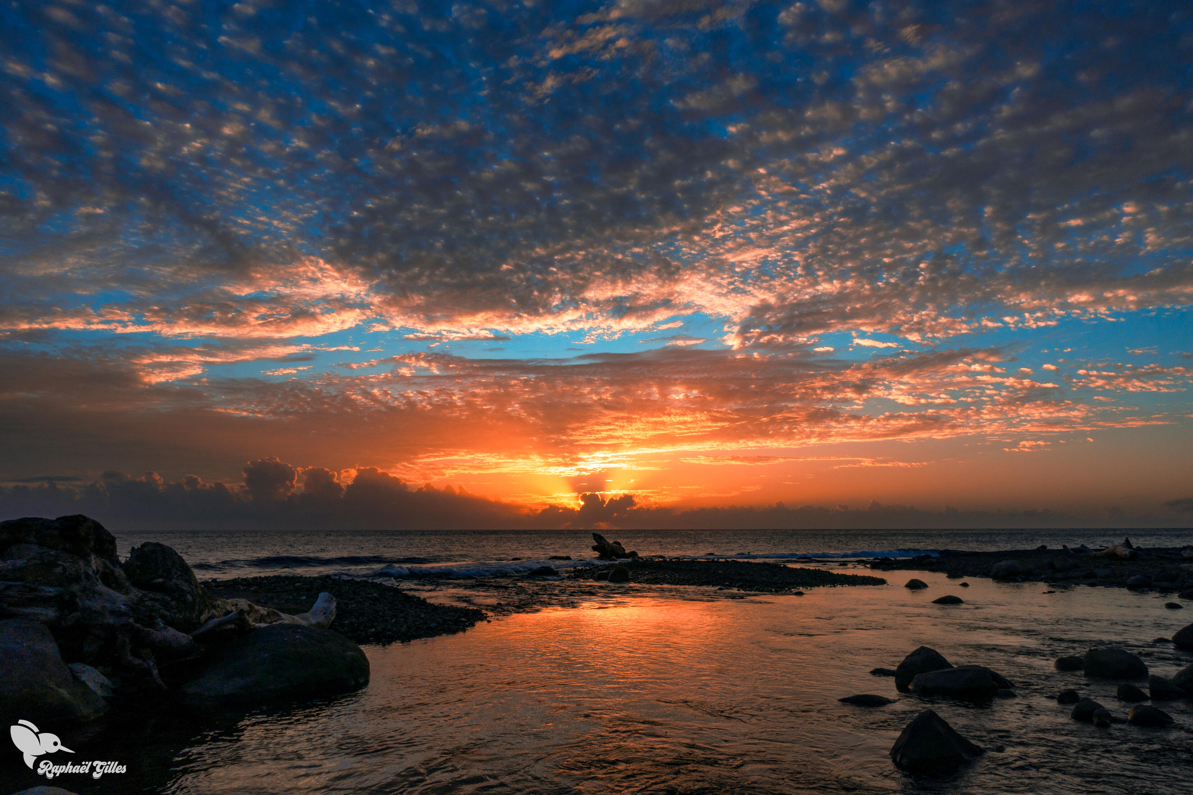 Un coucher de soleil sur la mer. Une rivière en premier plan se jette dans la mer.