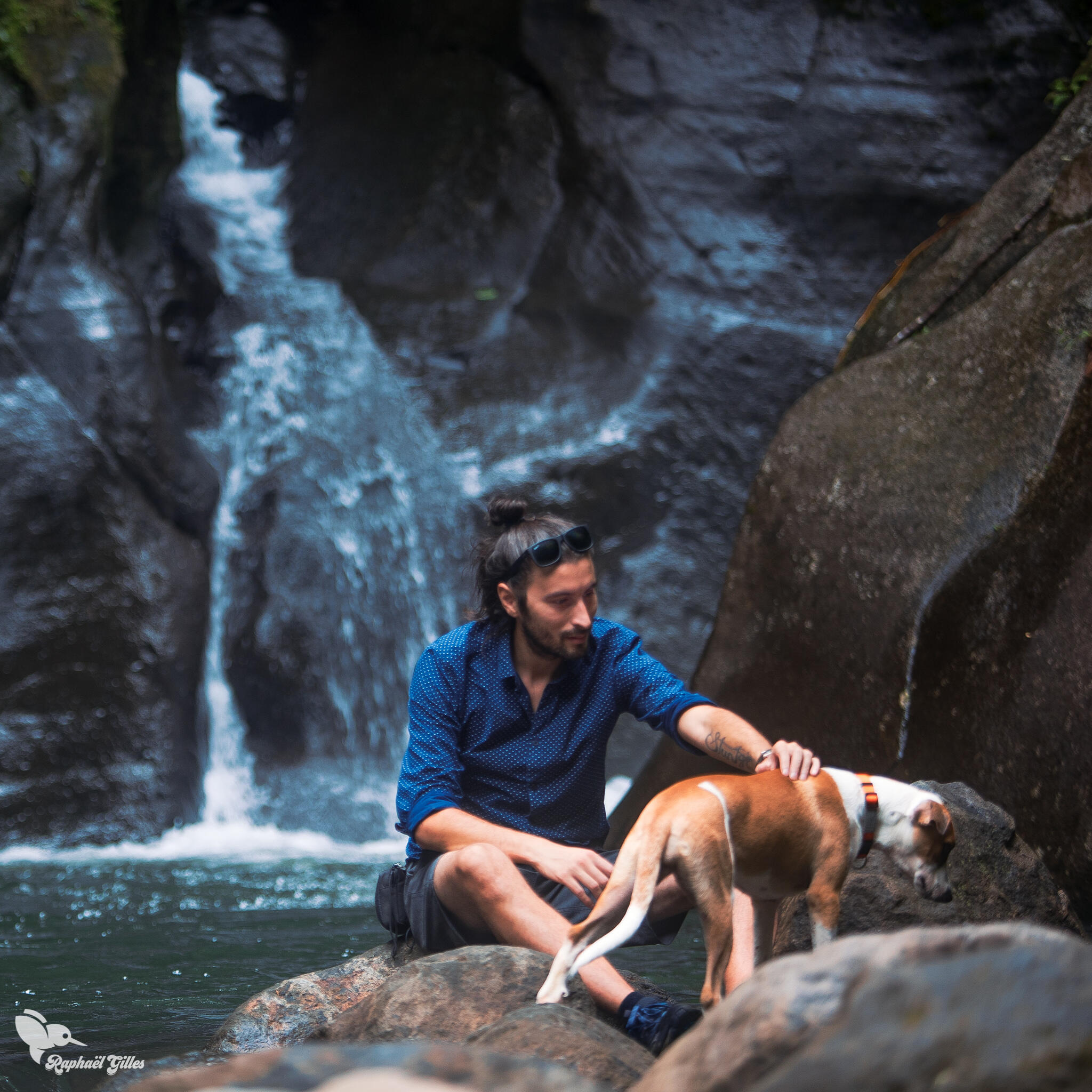 Un homme assis sur un sol rocheux au bord d'une cascade caresse un chien.