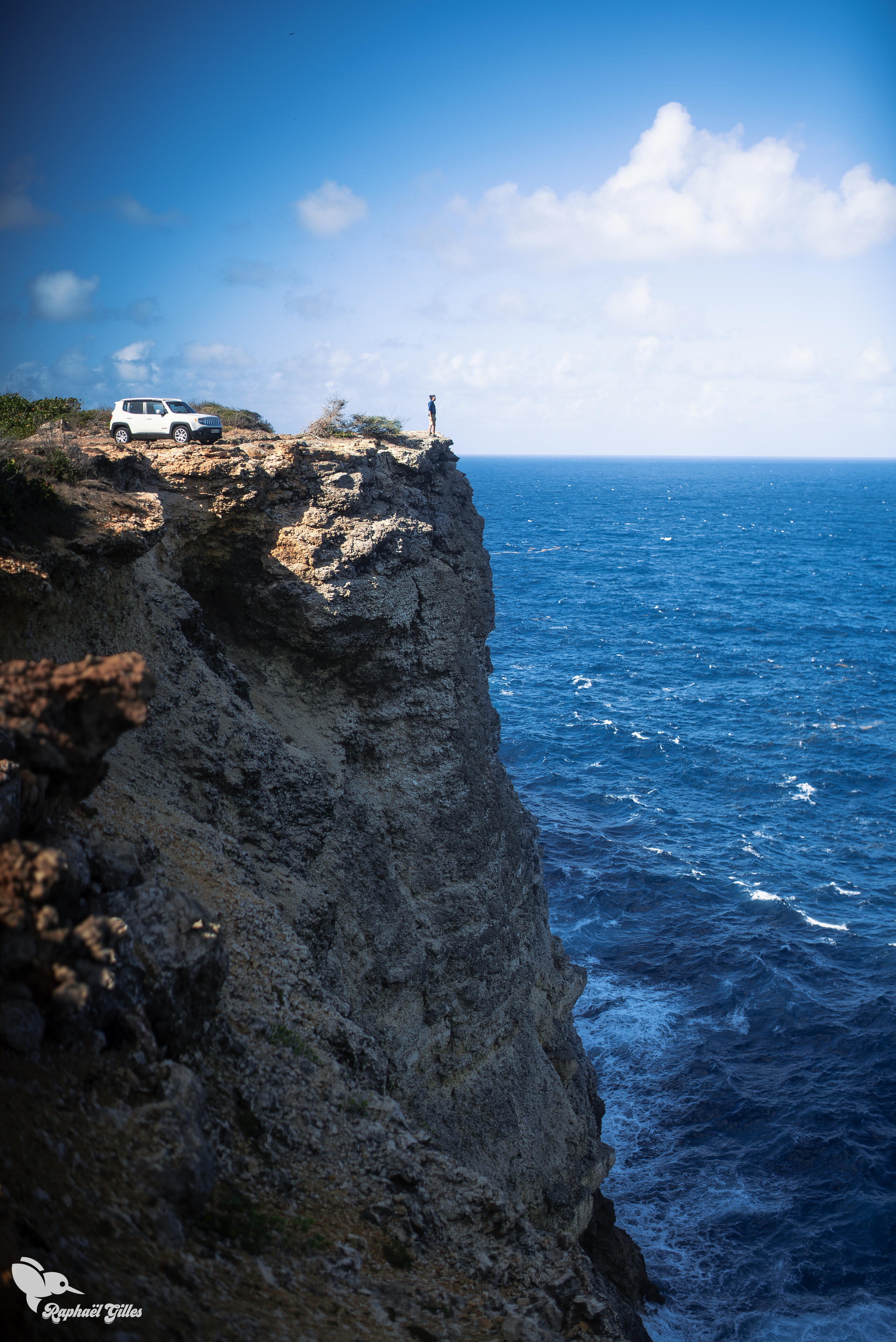 Un homme au bord d'une falaise vertigineuse.
Une voiture garée plus loin.