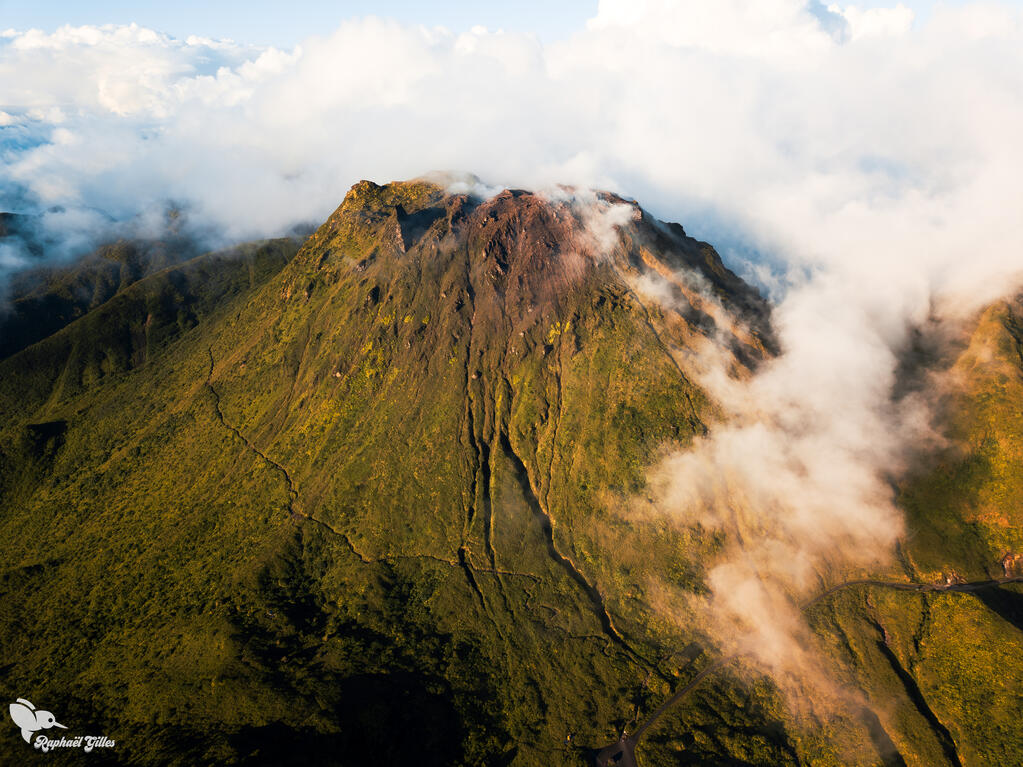 Photo prise au drone.
Un volcan (la Soufrière) dont le sommet est entouré de nuages. Des crevasses sur les flancs.