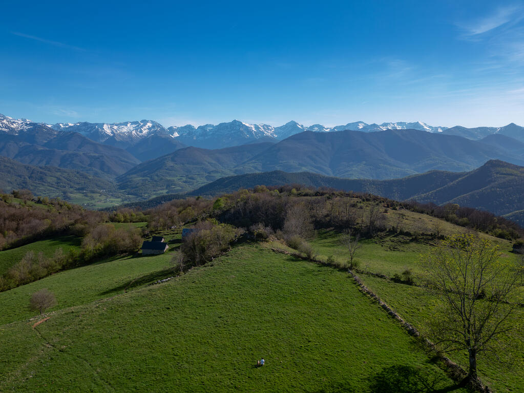 Photo prise au drone.
Une prairie au sommet d'une montagne en premier plan. Les Pyrénées ariégeoises enneigées à l'horizon.